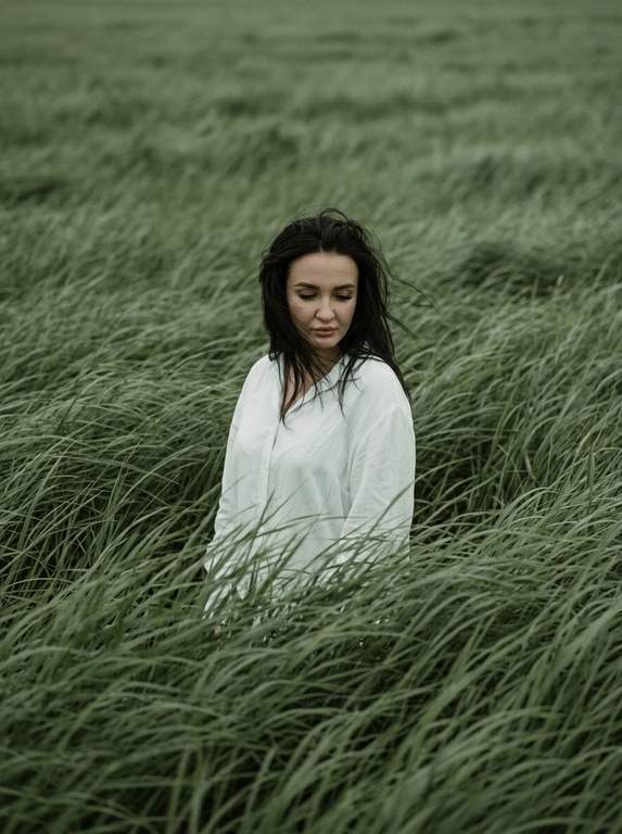 Create cinematic, atmospheric editorial portrait using the attached photo as the exact face and identity reference. A solitary model stands alone in the middle of vast wind-swept sage green grass fields. Tall grass surrounds the subject completely, flowing in strong currents like ocean waves, partially occurring the body did creduns mononaepin dna soutude. the subject wedis c simple, loose white shirt that softly contrasts with the rich green landscape hair is slightly messy, with natural strands moving in the wind. The head is gently tilted downward, eyes cast toward the ground, conveying a quiet introspective, melancholic mood. Expression is calm, distant, and emotionally restrained. Compose the frame slightly off-center, from a subtle elevated angle.
No visible horizon, the frame is fully immersed in grass texture. Use a muted natural palette of sage green, deep olive, and soft off-white. Shoot telephoto perspective, shallow depth of field, cinematic framing. Film photography destek with visible grain, soit edge tallon, atmospheric wind driven motion blur in the grass. Quiet, poetic storytelling: ultra-detail natural magazine-cover editorial quality. No text, no watermark.