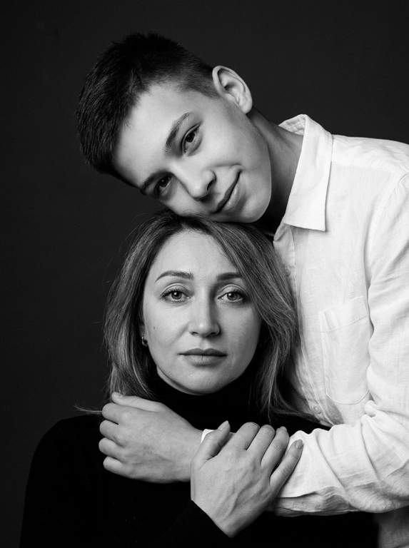 Родитель и ребёнок с внешностью как на приложенных фото, художественная семейная студийная фотосессия. Композиция — вертикальный портрет, крупный план лиц. Родитель внизу смотрит прямо в камеру, лицо фронтально. Ребёнок сверху, голова повернута набок, щека лежит на голове родителя, смотрит в камеру с нежной улыбкой. Рука ребёнка обнимает родителя за шею, видна в кадре, родитель держит руку ребёнка своей рукой.
Одежда: ребёнок в белой льняной рубашке, родитель в чёрной водолазке. 
Фон — тёмно-серый почти чёрный однотонный студийный, создаёт контраст. Освещение — мягкое боковое студийное, создаёт объём и глубину.
Чёрно-белая художественная фотография с выразительными контрастами. Атмосфера нежная, интимная, спокойная.