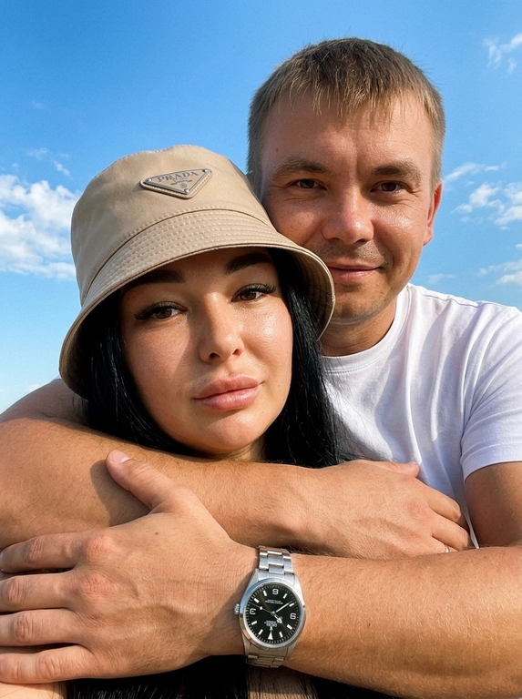 Кинематографичный кадр крупным планом (selfie style), пара в объятиях (классическое "обнимашки сзади") на фоне ярко-голубого неба с облаками.  Женщина на ней бежевая панама с брендовой надписью. Её кожа выглядит сияющей, подсвеченной мягким естественным солнечным светом. Выражение лица спокойное, глубокий взгляд, направленный прямо в камеру. Мужчина стоит строго позади девушки и обнимает её сзади, одна его рука обнимает её за плечи/шею спереди — предплечье лежит поперёк её ключиц, кисть свободно свисает с её плеча, на запястье классические серебряные часы с черным циферблатом. Мужчина в белой футболке, выражение лица доброжелательное, его взгляд прямо в объектив. Ракурс сверху, камера сфокусирована на лице девушки. Высокий контраст, насыщенные цвета, мягкая прорисовка деталей кожи. Эффект пленочной фотографии, легкое зерно, атмосфера летнего дня, эстетика социальных сетей. Телефона не видно в кадре.