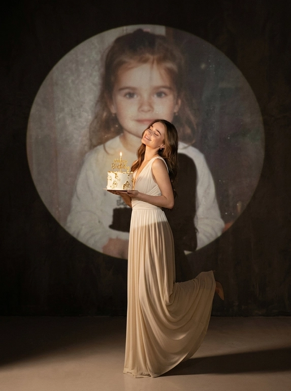 Create ultra-realistic cinematic birthday photoshoot, elegant studio with a dark background and a large circular spotlight projection on the wall. Inside the spotlight, a faded vintage childhood photo of the same girl(the reference image 2nd given)appears as a nostalgic memory, slightly transparent with old photo texture and soft blur. In the foreground, the present-day version of the same model (the reference image 1st given)  holding a white birthday cake decorated with gold accents with a “Happy Birthday". 

If the model is a woman, then she is wearing a cream long gown. If the model is a man, he is wearing a suit.

her head tilted back, eyes closed, natural smile,  cinematic shadow cast on the wall, warm soft spotlight lighting.

If the model was a woman, she additionally lifted her leg in an elegant pose.
