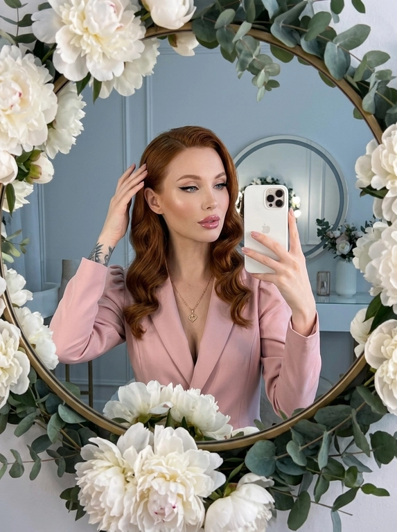 Максимально реалистичное селфи в зеркале в эстетике luxury soft glam. Модель стоит перед круглым зеркалом с изящной рамой, украшенной белыми живыми пионами. Цветы обрамляют зеркало нежно и дорого, создавая ощущение утончённого, премиального кадра. Композиция чистая и визуально собранная: модель находится в центре отражения, кадр выглядит эстетично, дорого и современно.

Поза естественная и уверенная: одной рукой модель держит современный смартфон, второй рукой слегка касается волос, создавая живой и непринуждённый жест. Селфи выглядит не постановочным, а как красивый спонтанный кадр из дорогой life-style съёмки.

Макияж — выразительный soft glam: чёткие чёрные стрелки, длинные пышные ресницы с лисьим эффектом, внутренние уголки глаз подсвечены сияющими тенями. Кожа ровная, ухоженная, с мягким влажным сиянием. Губы подчёркнуты карандашом и покрыты полупрозрачным глянцевым блеском, создающим аккуратный объём.

На модели ее образ выглядит женственно, современно и дорого. 

Смартфон современный, в минималистичном белом матовом чехле. Если нужен более дорогой вайб, без лишнего декора и ярких деталей.

Освещение холодное, мягкое и чистое, как в дорогой beauty-съёмке или premium dressing room. Свет красиво подчеркивает сияние кожи, текстуру ткани, свежесть пионов и блеск украшения. Атмосфера — роскошная, женственная, ухоженная, без дешёвого глянца.

Фотореализм максимальный: высокая детализация кожи, ткани, цветов, аксессуаров и отражения в зеркале. Лёгкий цифровой шум, мягкая резкость, деликатная зернистость, естественная глубина кадра, высокая контрастность, эффект живого фото, без CGI и без искусственного AI-эффекта.

Какая будет прическ?: крупные голливудские волны, мягкий блеск волос, аккуратная укладка на одну сторону.

Какой будет образ на фото? : Элегантный пудрово-розовый жакет с акцентными рукавами-фонариками и глубоким V-образным вырезом. На шее — тонкая изящная цепочка с ювелирным кулоном в форме сердца.