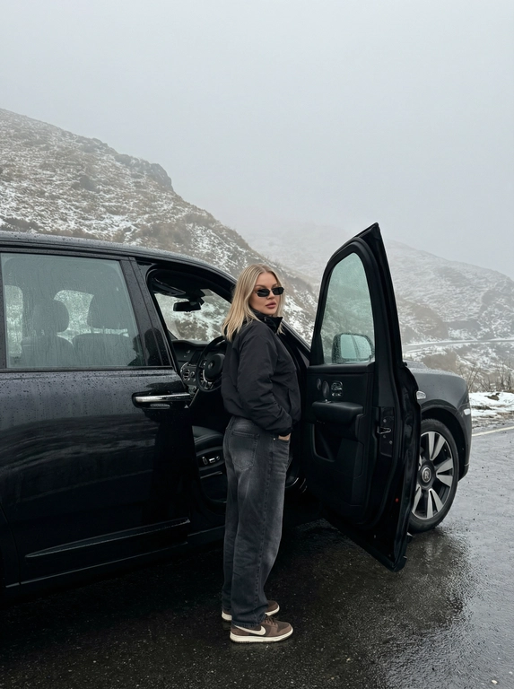 Не менять черты лица человека с исходного фото. Человек рядом с стоит рядом с машиной, машина на горной дороге, дорога мокрая от тумана, машина в каплях от тумана. Чёрный анорак Planta Rosa, маленькие чёрные солнцезащитные очки без оправы, мешковатые серо-чёрные джинсы с низкой посадкой, длинные, коричневые замшевые кроссовки Nike Dunk, руки в карманах. Машина без номеров. Фон — гиперреалистичные заснеженные холмы Мегхалайи, без зелени, туманные горы, пасмурный мягкий дневной свет, без размытия, без изменения лица или позы, без дополнительных объектов. Фото снято на iPhone 17 Pro, без фильтров на лице, без ретуши, перспектива снизу, формат 9:16, машина как на исходном фото, фон не размывать, притемнённая экспозиция, открывает дверь машины, стоит спиной, смотрит в камеру, будто сейчас сядет за руль.


Какая будет машина?: Стоит рядом с машиной Rolls-Royce Cullinan