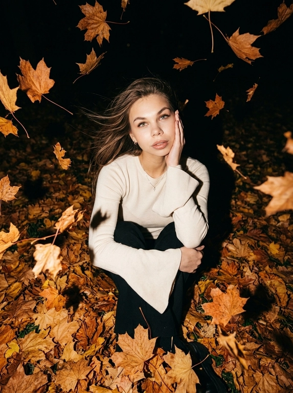 Ultra-realistic candid NIGHT autumn photo. The woman sits curled up directly in a thick carpet of dry autumn maple leaves, knees pulled toward her chest. Camera positioned very high above her, almost directly overhead, dramatic top-down perspective. She raises her face toward the camera, one hand gently supporting her cheek, the other arm resting across her knees.
She wears a fitted white knitted top with very long dramatic FLARED BELL SLEEVES, fitted through the arms and strongly widening from the elbows.
Strong autumn wind blows her hair.

It is DARK outside, almost nighttime. The background beyond the flash falls into deep natural darkness. Shot with a powerful direct on-camera flash: her face, white sweater and the closest leaves are brightly illuminated, while distant leaves rapidly disappear into black shadows. Dozens of dry orange, rust and brown leaves are FLYING THROUGH THE AIR around her, caught mid-flight by the flash; some pass very close to the lens and create strong realistic motion blur and streaks. A few strands of hair also have motion blur from the wind. Face remains sharp. Raw spontaneous flash photography, high contrast, deep blacks, warm saturated autumn tones, slight grain, imperfect handheld framing, realistic textures, no studio lighting, no HDR, no AI look.