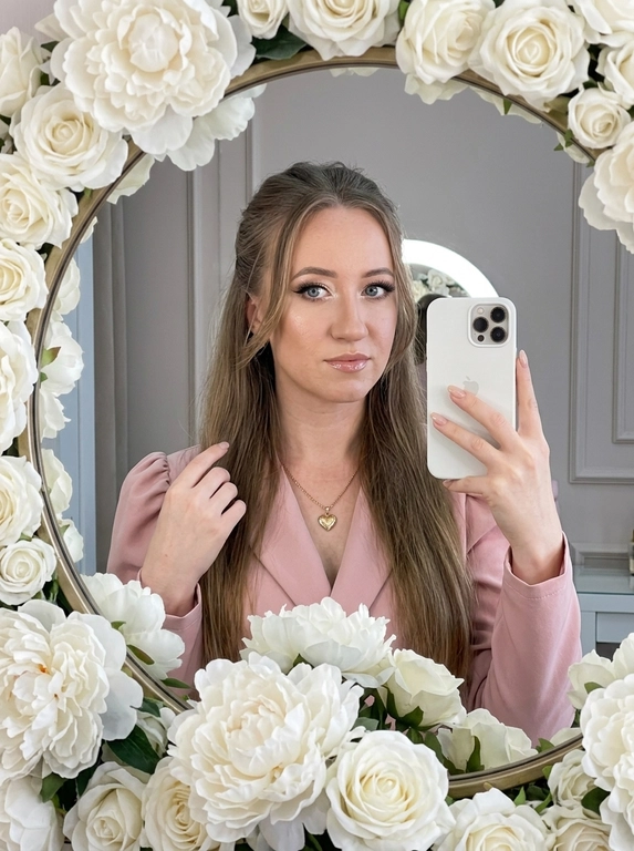Максимально реалистичное селфи в зеркале в эстетике luxury soft glam. Модель стоит перед круглым зеркалом с изящной рамой, украшенной белыми живыми пионами. Цветы обрамляют зеркало нежно и дорого, создавая ощущение утончённого, премиального кадра. Композиция чистая и визуально собранная: модель находится в центре отражения, кадр выглядит эстетично, дорого и современно.

Поза естественная и уверенная: одной рукой модель держит современный смартфон, второй рукой слегка касается волос, создавая живой и непринуждённый жест. Селфи выглядит не постановочным, а как красивый спонтанный кадр из дорогой life-style съёмки.

Макияж — выразительный soft glam: чёткие чёрные стрелки, длинные пышные ресницы с лисьим эффектом, внутренние уголки глаз подсвечены сияющими тенями. Кожа ровная, ухоженная, с мягким влажным сиянием. Губы подчёркнуты карандашом и покрыты полупрозрачным глянцевым блеском, создающим аккуратный объём.

На модели ее образ выглядит женственно, современно и дорого. 

Смартфон современный, в минималистичном белом матовом чехле. Если нужен более дорогой вайб, без лишнего декора и ярких деталей.

Освещение холодное, мягкое и чистое, как в дорогой beauty-съёмке или premium dressing room. Свет красиво подчеркивает сияние кожи, текстуру ткани, свежесть пионов и блеск украшения. Атмосфера — роскошная, женственная, ухоженная, без дешёвого глянца.

Фотореализм максимальный: высокая детализация кожи, ткани, цветов, аксессуаров и отражения в зеркале. Лёгкий цифровой шум, мягкая резкость, деликатная зернистость, естественная глубина кадра, высокая контрастность, эффект живого фото, без CGI и без искусственного AI-эффекта.

Какая будет прическ?: полусобранная укладка, верхняя часть волос аккуратно убрана назад, мягкие пряди обрамляют лицо.

Какой будет образ на фото? : Элегантный пудрово-розовый жакет с акцентными рукавами-фонариками и глубоким V-образным вырезом. На шее — тонкая изящная цепочка с ювелирным кулоном в форме сердца.