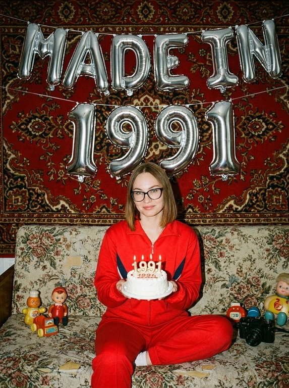 Создай крупный портрет, не меняя черты лица. На фотографии модель в стиле «лихие 90-е» в России. Модель сидит на старом диване с цветочной обивкой на фоне красного советского ковра на стене и смотрит в камеру с лёгкой улыбкой. На ковре висят большие надувные серебряные фольгированные шары в форме цифр и надпись «MADE IN». На модели классический красный спортивный костюм того времени и кроссовки. В руках маленький праздничный белый торт со свечами. Рядом на диване лежат игрушки: советские игрушки и старая советская кукла. Фото со вспышкой. Освещение мягкое, создающее атмосферу ностальгии. Высокое разрешение, реализм. Модель в олдскульном спортивном костюме сидит расслабленно, с лёгкой улыбкой. Ретро-вайб, приглушённые цвета, зернистость плёнки, детализированные текстуры ткани и шерстяного ковра.

Какая будет цифра?: Цифра 1991
