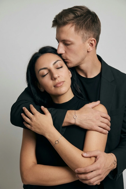 Мужчина и женщина позируют в студии в объятиях. Мужчина стоит позади женщины, обнимая её обеими руками; его тело повернуто к ней, голова слегка наклонена вниз, губы касаются её лба. Женщина немного отклоняет голову назад к его плечу, её глаза закрыты. Кадр — средний, от талии до головы, снят на уровне глаз. Композиция плотная, акцентирует линии рук и соприкосновение лиц. Женщина стоит, прямо обращённая к камере.
На ней облегающий чёрный топ без рукавов с высоким воротом, из плотной матовой ткани.
Мужчина одет в чёрную футболку и чёрную неброскую куртку. На обоих запястьях — тонкие металлические браслеты с минималистичными круглыми подвесками, а женщина носит небольшие светлые металлические серьги с гладкой поверхностью.
Обстановка минималистичная: однотонный светло-серый фон без текстуры.
Освещение — мягкий студийный свет: рассеянный фронтальный и боковой свет создаёт плавные переходы света и тени на лицах и руках. Тени мягкие, средней глубины. Контраст умеренный, без жёстких бликов. Свет равномерно распределён по фигурам, фон подсвечен мягко, без выраженных градиентов.
Съёмка на портретный объектив 85 мм, малая глубина резкости с чётким фокусом на лицах и руках; фон слегка размыт, текстуры кожи и ткани высоко детализированы. Это минималистичный студийный портрет, высокодетализированный и фотореалистичный.
Минималистичное романтическое студийное настроение, нейтральные тёплые тона кожи, низкая–средняя насыщенность, мягкое рассеянное освещение, средний контраст с нежными тенями, плавные переходы бликов, естественная передача кожи, чистый светлый фон, тонкий тональный баланс, современная мягкая редакционная цветокоррекция.

Не изменять мою прическу