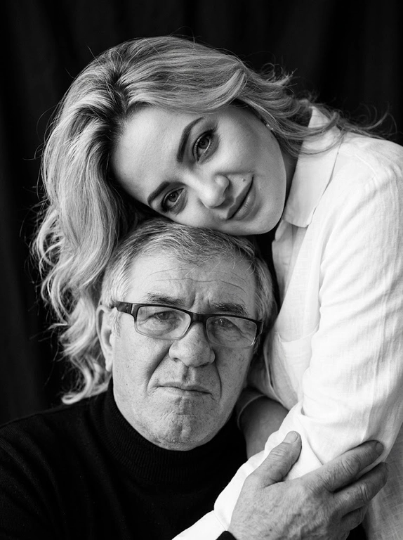 Родитель и ребёнок с внешностью как на приложенных фото, художественная семейная студийная фотосессия. Композиция — вертикальный портрет, крупный план лиц. Родитель внизу смотрит прямо в камеру, лицо фронтально. Ребёнок сверху, голова повернута набок, щека лежит на голове родителя, смотрит в камеру с нежной улыбкой. Рука ребёнка обнимает родителя за шею, видна в кадре, родитель держит руку ребёнка своей рукой.
Одежда: ребёнок в белой льняной рубашке, родитель в чёрной водолазке. 
Фон — тёмно-серый почти чёрный однотонный студийный, создаёт контраст. Освещение — мягкое боковое студийное, создаёт объём и глубину.
Чёрно-белая художественная фотография с выразительными контрастами. Атмосфера нежная, интимная, спокойная.