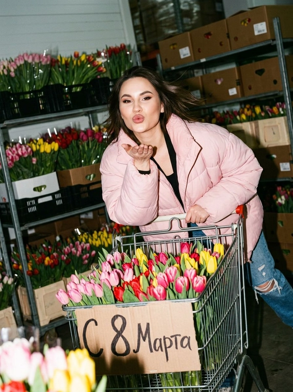 Создай фото, не меняя черты лица. Женщина толкает тележку, переполненную цветов. На тележке табличка «С 8 Марта». На ней большой нежно-розовый пуховик, расстёгнутый, голубые джинсы с потертостями, топ. Безупречный макияж. Она наклоняется вперёд над ручкой тележки с игривым выражением лица, словно посылая поцелуй в сторону камеры. За ней — полки, заставленные теми же цветами. Непринуждённый, естественный, спонтанный стиль фотографии. Съёмка с резкой прямой вспышкой — сильные тени, пересвеченные участки, неравномерная экспозиция. Наклонная композиция: объекты не по центру, обрезанные конечности, неидеальное выравнивание. Размытие в движении, неидеальная фокусировка, съёмка в середине движения или в середине выражения эмоций. Шум и зернистость — без ретуши или сглаживания. Небрежная, интимная, реалистичная, документальная атмосфера.


Какие будут цветы?: Тележка полная тюльпанов. Полки заставленные тюльпанами.