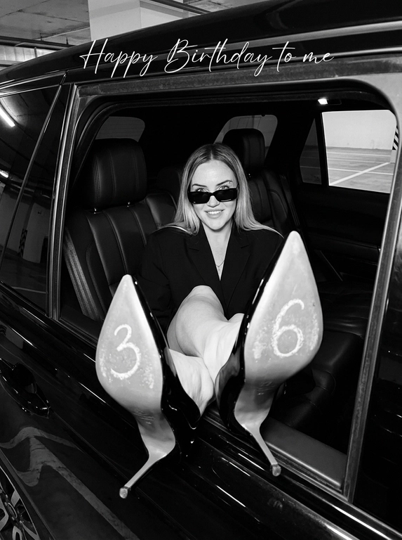 Фотореалистичная чёрно-белая fashion-фотография. Съёмка снаружи через открытое боковое окно чёрного премиального внедорожника в подземном паркинге. Женщина в узких прямоугольных чёрных солнцезащитных очках и объёмном чёрном жакете, расслабленно сидит в кожаном кресле и смотрит в камеру с лёгкой сомкнутой улыбкой. Обе ноги подняты к открытому окну, подошвы чёрных лаковых туфель острыми носами и тонкими высокими каблуками направлены прямо в объектив и занимают большую часть нижней половины кадра; лицо находится глубже, справа. Верх туфель — глянцевый чёрный лак с контрастными бликами, а вся обращённая к камере поверхность подошв — равномерно матовая, серого цвета, немного светлее верха, с тонкой естественной фактурой и мягким рассеиванием света; серый цвет является цветом материала и сохраняется по всей длине подошвы, включая узкую часть под сводом стопы, без глянца, зеркальных отражений, металлического блеска, ярких бликов и белых засветов. На левой для зрителя подошве белым мелом крупная цифра «», на правой — «»; цифры хорошо читаются, с естественно неровными краями и пудровой фактурой мела. Выраженная перспектива с сильным эффектом переднего плана. Прямая вспышка, глубокие тени, контрастные блики только на лаковой поверхности туфель и кузове автомобиля, подошвы освещены ровно и матово. Лёгкая плёночная зернистость, реалистичная анатомия и натуральная текстура кожи. Атмосфера дерзкой, стильной birthday-съёмки. В верхней трети кадра крупная тонкая белая рукописная надпись «Happy Birthday to me». Без интерфейса, значков воспроизведения, водяных знаков, лишних конечностей и анатомических артефактов.

Цифры на каблуках: На левом "3"  На правом "6"

Доп. характеристики: Пирсинг в брови, канапушки по телу и лицу, волосы прямые