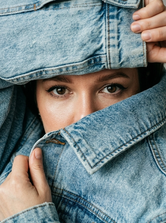 Экстремально крупный фотореалистичный fashion editorial портрет. Камера очень близко к лицу, плотный кадр, ракурс почти на уровне глаз с лёгким верхним углом. Модель не стоит прямо, а будто прячется внутри объёмной светло-голубой джинсовой куртки. Поза читается только через руки, лицо и ткань.

Весь кадр заполнен джинсой и руками. Сверху большой слой денима нависает над бровями, снизу поднятый воротник диагонально закрывает лицо до переносицы. Видны только глаза, брови, переносица и небольшой участок кожи вокруг глаз. Рот, подбородок, щёки, шея и причёска скрыты. Волосы почти не видны, максимум лёгкая тень у линии роста волос.

Одна рука сверху справа держит верхний край джинсовой куртки, частично обрезана кадром. Вторая рука снизу слева подтягивает воротник ближе к лицу. Пальцы естественно сжимают плотную ткань, создавая натяжение, складки и объём. Композиция тесная, асимметричная, с диагональными линиями ткани вокруг глаз.

Деним главный элемент кадра: светло-голубая washed denim ткань, плотная, матовая, тяжёлая, с выраженной фактурой, видимым плетением, мягкими потёртостями, заломами, массивными швами и контрастной строчкой. Джинсовая куртка выглядит натурально, объёмно и очень близко к камере.

Глаза смотрят прямо в камеру из-под бровей. Взгляд спокойный, глубокий, уверенный и слегка загадочный. Один глаз немного в тени, второй более чёткий и освещённый. Макияж глаз аккуратный: тонкая вытянутая стрелка, лёгкий cat-eye эффект, подчёркнутые ресницы, натуральные брови. В глазах живые естественные блики.

Кожа вокруг глаз реалистичная, с натуральной текстурой, мягкими порами и естественными бликами. Без пластикового сглаживания, без чрезмерной ретуши, без AI-эффекта.

Освещение мягкое, естественное, слегка контрастное, как в дорогой editorial-съёмке. Свет подчёркивает глаза, переносицу, руки, швы и складки денима. Тени глубокие, но натуральные. Цвета спокойные и реалистичные: светло-голубой деним, нейтральный тон кожи, мягкие тени.

Фон полностью отсутствует. В кадре только джинсовая ткань, руки и узкий фрагмент лица. Ultra realistic close-up, luxury denim editorial, natural film grain, shallow depth of field, soft sharpness, high detail, realistic skin texture, realistic denim texture, no CGI, no plastic skin, no over-retouching.

Negative prompt: visible hairstyle, loose hair, open face, full face, mouth visible, chin visible, neck visible, full body, standing pose, normal jacket pose, symmetrical framing, empty background, studio backdrop, glossy denim, plastic skin, AI smoothing.
