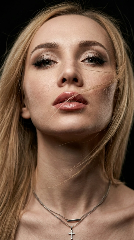 Фотореалистичный цветной beauty-портрет, экстремально крупный план (лицо + шея + ключицы), низкий ракурс снизу вверх, голова слегка запрокинута. Волосы хаотично пересекают лицо, несколько прядей на губах и щеке, ощущение ветра и движения. Взгляд открытый, дерзкий.
Макияж: дымчатые тени с мягким светлым шиммером, чёткая чёрная стрелка, густые ресницы, аккуратно оформленные брови. Губы, с влажным глянцевым блеском, чёткий контур. Кожа без ретуши — естественный чистый  тон, живая текстура, видимые поры, мягкие цветовые переходы света и тени.Модадвя девушка не меняй лицо 
Фон полностью чёрный, минимализм.
Свет: один боковой soft key light + лёгкий fill, высокий контраст, глубокие тени, выразительный рельеф скул и губ. Кинематографичный цветовой свет с естественными оттенками кожи и лёгкими тёпло-холодными нюансами. Лёгкий rim/back light для отделения от фона.
Съёмка в стиле fashion editorial. Сверхрезкий фокус на глазах и губах, малая глубина резкости. 
Ideal quality settings: ultra-realistic 8K, hyper-detailed, sharp focus; shallow depth of field, natural bokeh; HDR ,; cinematic color grading; lens 85mm / 50mm / 35mm; aperture f/1.4–f/2.8; key light at 45°, soft shadows; clean white balance; natural skin tones, glossy highlights.