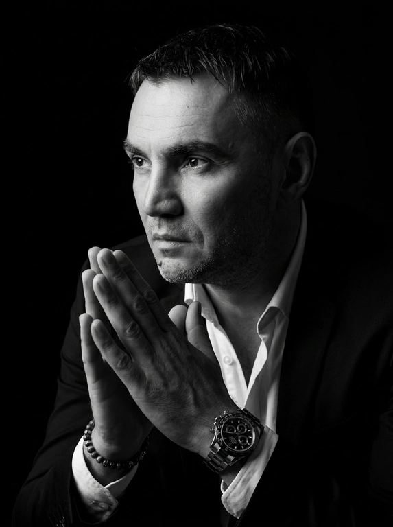 A hyper-realistic 8K cinematic portrait of a uploaded person,
captured in a dramatic Chiaroscuro style. The subject is seated at a
three-quarter angle, leaning slightly forward with a relaxed yet
commanding posture. His face is turned away from the lens, with
one side of his face clearly illuminated while the other is
completely lost in deep, velvety black shadows. His expression is
contemplative, not looking at the camera. The pose is precise: his
hands are clasped together near his chest; one hand is held
straight with fingers slightly tilted, while the other hand is
positioned so only the fingers are visible, resting over the first. He
wears a luxury black chronograph watch with a detailed metal link
bracelet, and a beaded bracelet. His attire is a sharp
black suit jacket over a white dress shirt with the top buttons
open, showing refined fabric textures. The background is a solid,
seamless black. The lighting is sharp and directional, highlighting
the intricate details of his hair,
the moisture in his eyes, and the glint off the watch face.
Cinematic black-and-white color grade with rich contrast and
smooth, natural skin transitions. Shot on 85mm lens, f/1.8, high-
resolution commercial photography