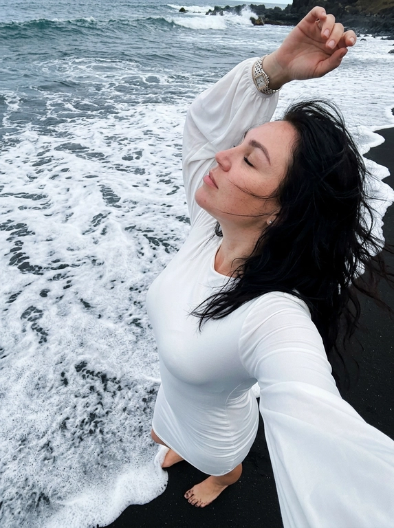 Снято на iPhone, селфи с руки, телефона строго в кадре нет.

Женщина стоит у самой кромки моря на чёрном вулканическом песке. Камера удерживается высоко над головой одной рукой и направлена строго вниз под углом, создавая эффект динамичного селфи сверху. Она находится в центре кадра, верхняя часть тела занимает большую часть фотографии.

Поза: голова слегка запрокинута назад, лицо повернуто в профиль к солнцу, глаза закрыты, губы расслаблены. Корпус слегка изогнут, создавая красивую линию талии. Волосы развеваются морским ветром.

Одежда: белое облегающее платье с длинными свободными рукавами в стиле Skims. Маникюр короткий, квадратной формы, френч. Кожа слегка загорелая.

Украшения: тонкая цепочка с небольшим крестиком, серьги-крестики, часы Cartier Panthère из стали.

Обстановка: чёрный вулканический пляж, белая морская пена накатывает прямо к ногам и краю платья, контрастируя с тёмным песком. Волны образуют красивую изогнутую линию вдоль кадра. Яркий солнечный день, людей вокруг нет.

Освещение: естественный яркий дневной свет, солнце высоко над горизонтом, мягкие естественные тени, насыщенные цвета моря, белой пены и чёрного песка. Без вспышки.

Стиль: живое атмосферное travel/lifestyle-фото, ощущение случайно пойманного момента, натуральная текстура кожи, лёгкое плёночное зерно, без фильтров, не размывай фон, без текста, без водяных знаков.