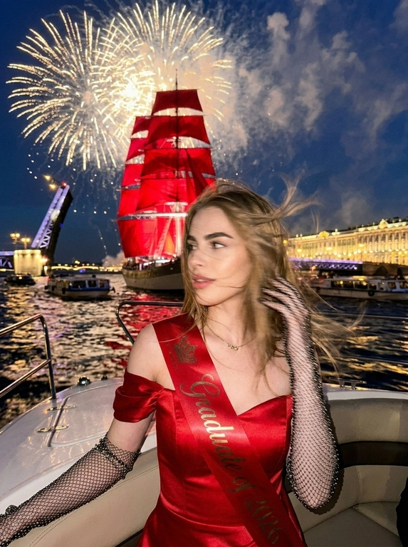Создай портрет. Ночной экстремально близкий портрет в стиле дорогого fashion-снимка, крупный план, ощущение случайно пойманного момента. Модель, сидящая в энергичной позе на роскошной лодке, плывущей по каналам Санкт-Петербурга во время праздника - "Алые паруса". Волосы развиваются от ветра, слегка на лице. Шикарный вечерний макияж, у нее еле заметная улыбка. 

На заднем плане воды канала, отражающие на горизонте появляются исторические здания, характерные для Санкт-петербурга, рядом с лодкой плывет огромное количество парусных лодок, маленьких яхт, туристических лодок и небольшие лодки вдалеке. Интерьер лодки изысканный. На фоне разводной мост и в центре внимания огромный известный корабль с алыми парусами, паруса подсвечены, с корабля красиво бью прожектора, он в центре внимания, выглядит величественно. Здания подсвечиваются. Все небо залито огромным количеством салютов и фейерверков, а так же светом прожекторов, салюты во весь кадр

Кадр намеренно неидеальный: сильный motion blur, длинная выдержка, высокий ISO, выраженное зерно. Жёсткий контраст, кинематографичная небрежность, ощущение ночи, роскоши и живого, спонтанного кадра. Атмосфера праздника, величественного события. Модель сидит грациозно прямо к кадру, голова повернута в сторону, лицо немного размазано из-за движения, девушка в расфокусе, не смотрит в кадр. Весь фокус на корабле с алыми парусами, они четко подсвечены.

Атласная красная лента "Выпускница 2026"?: На ней атласная красная лента "Выпускница 2026"

Какое будет платье?: Она одета в черное очень приталенное атласное черное платье ниже колена с открытыми плечами, на ней длинные перчатки из сетки со стразами; золотые сверкающие серьги, тонкая золотая цепочка-ожерелье.
