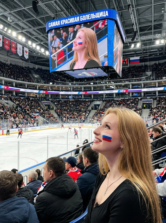 Используй загруженное фото как точный референс лица. Сохрани ту же личность, ту же структуру лица, ту же форму глаз, тот же нос, те же губы, тот же тон кожи, тот же цвет и длину волос. Не изменяй черты лица. Сохрани пропорции. Ультрареалистичная фотография внутри большой крытой хоккейной арены во время живого матча. Передний план: та же женщина-болельщица сидит на трибунах, профиль, смотрит вверх на табло арены с мягкой эмоциональной улыбкой. Она сидит среди зрителей. На щеке аквагрим в виде российского флага (белая, синяя, красная полосы). Естественный макияж. Реалистичная текстура кожи. Над ней — огромное центральное табло арены, подвешенное под потолком. На экране табло её лицо показано в прямой трансляции с того же ракурса. Позади неё на изображении табло чётко видны трибуны и места арены. На заднем плане виден ледовый каток. ВАЖНО: вся сцена в фокусе. Глубокая глубина резкости. Чёткий задний план. Без размытой толпы. Без малой глубины резкости. Всё чётко и детализировано. Освещение: реалистичное освещение арены, слегка холодные тона. Текст на табло: «САМАЯ КРАСИВАЯ БОЛЕЛЬЩИЦА». Снято на iPhone 17 Pro. Естественная съёмка на смартфон. Без стилизации. Без AI-эффекта. Высокий реализм. Вид как при диафрагме f/8, документальная спортивная фотография, высокая чёткость по всему кадру.