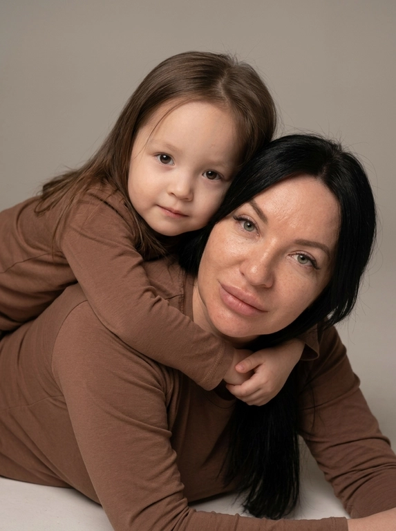 Создай студийный портрет родителя и ребёнка, максимально точно сохранив черты лица и выражения с референсных изображений. Не изменяй и не искажай лица, воспроизведи их с фотографической точностью.

Композиция:

Крупный план, ракурс на уровне глаз.

Родитель и ребёнок лежат на животе, расположены боком к камере.

Ребёнок лежит на спине родителя и мягко обнимает его за плечи.

Оба смотрят прямо в камеру, создавая тёплый и близкий зрительный контакт.

На родителе и ребёнке одинаковые коричневые лонгсливы без принтов и декора.

Естественный внешний вид, ровный тон кожи.

Мягкое студийное освещение, спокойная семейная атмосфера, натуральная текстура кожи, высокая детализация, максимальный фотореализм. Без ламп в кадре и прочего декора.

Формат 9:16.

Качество 8K.