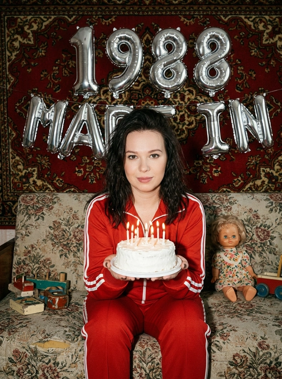 Создай крупный портрет, не меняя черты лица. На фотографии модель в стиле «лихие 90-е» в России. Модель сидит на старом диване с цветочной обивкой на фоне красного советского ковра на стене и смотрит в камеру с лёгкой улыбкой. На ковре висят большие надувные серебряные фольгированные шары в форме цифр и надпись «MADE IN». На модели классический красный спортивный костюм того времени и кроссовки. В руках маленький праздничный белый торт со свечами. Рядом на диване лежат игрушки: советские игрушки и старая советская кукла. Фото со вспышкой. Освещение мягкое, создающее атмосферу ностальгии. Высокое разрешение, реализм. Модель в олдскульном спортивном костюме сидит расслабленно, с лёгкой улыбкой. Ретро-вайб, приглушённые цвета, зернистость плёнки, детализированные текстуры ткани и шерстяного ковра.

Какая будет цифра?: Цифра 1988
Девушка в спортивном костюме