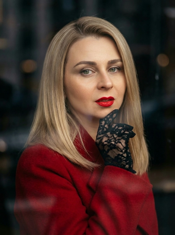 Фотореалистичный fashion-портрет женщины.
Элегантная, уверенная, кинематографичная съемка в стиле high fashion editorial.
Крупный портрет по грудь, легкий поворот корпуса, взгляд прямо в камеру, слегка приоткрытые ярко-красные губы, выразительный макияж с черной стрелкой eyeliner.
На ней ярко-красное пальто с плотной тканью и аккуратным кроем, образ роскошный, дерзкий и женственный.
Одна рука в черной кружевной перчатке поднята к лицу, пальцы слегка касаются подбородка и губ, создавая изысканную позу.
Украшения минимальные, небольшие элегантные серьги.
Съемка через стекло или окно, на переднем плане мягкие размытые отражения и легкие полосы стекла, как у уличной модной фотографии.
Фон темный, размытый, с мягким боке и атмосферой города, но без отвлекающих деталей.
Освещение мягкое, теплое, контрастное, с красивыми тенями на лице, skin glow, естественная текстура кожи, дорогой журнальный стиль.
Цветокоррекция: теплые глубокие оттенки, акцент на красный цвет пальто и губ, благородные темные тени, cinematic mood.
Фото выглядит как дорогая съёмка для модного журнала, ultra realistic, high detail, sharp focus on face, shallow depth of field, 85mm portrait lens, soft film grain, luxury editorial aesthetics.
Негативный промпт
искаженное лицо, другая личность, лишние пальцы, лишние руки, деформированные губы, кривые глаза, плохая анатомия, мультяшность, пластик, чрезмерная ретушь, низкое качество, размытое лицо, шум, пересвет, артефакты, лишние украшения, лишний фон, неестественная кожа, плохой макияж, кривой нос, неправильные пропорции, двойное лицо

Какая будет укладка?: Укладка в стиле ретро-glam: волосы аккуратно уложены в крупные гладкие волны с выраженным объёмом у корней, мягким боковым пробором и глянцевой, ухоженной текстурой. Волны обрамляют лицо и создают элегантный образ в духе классической голливудской съёмки.