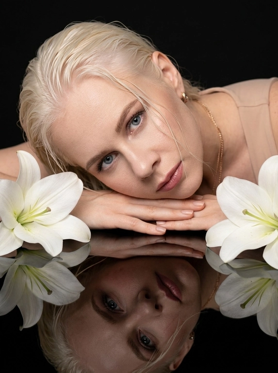 Фотореалистичный студийный beauty-портрет в эстетике дорогой рекламной кампании люксового косметического бренда. Минималистичная студия с глубоким матовым чёрным фоном. Перед моделью идеально гладкий чёрный зеркальный стол с чистым отражением лица, рук и цветов.

Женщина спокойно лежит на животе, корпус расположен немного по диагонали. Плечи расслаблены. Лицо находится почти вплотную к поверхности стола. Голова слегка наклонена вправо и мягко опирается щекой на сложенные кисти рук. Правая кисть лежит под щекой, левая свободно расположена сверху. Руки направлены по диагонали вдоль нижней части лица, пальцы расслабленные и естественно вытянутые. Лицо практически параллельно поверхности стола. Взгляд направлен прямо в объектив. Выражение спокойное, уверенное и слегка отстранённое. Губы мягко расслаблены.

В одном ухе небольшая изящная серьга с прозрачными бриллиантами. На модели минималистичный топ телесного оттенка с тонкими бретелями, почти полностью скрытый кадрированием.

Макияж выполнен в стиле luxury beauty campaign. Идеально ровный естественный тон кожи с деликатным сатиновым финишем. Лёгкий контуринг мягко подчёркивает скулы и овал лица. Нежные бежево-тауповые тени с плавной растушёвкой создают выразительную глубину взгляда. Верхнее межресничное пространство аккуратно затемнено, тонкая мягкая стрелка практически незаметна. Длинные, густые, идеально разделённые ресницы с объёмной премиальной тушью. Брови натуральные, аккуратно уложенные, с мягкой архитектурой и естественной густотой. На внутренних уголках глаз едва заметное сияние. Скулы подчёркнуты деликатным сатиновым хайлайтером без крупных блёсток. Губы объёмные, идеально очерченные пыльно-розовым карандашом, покрыты полупрозрачной розово-бежевой помадой с дорогим увлажняющим глянцевым финишем.

По обе стороны лица расположены две крупные полностью раскрытые белые лилии. Цветы находятся очень близко к объективу, частично выходят за границы кадра и создают симметричное обрамление лица. Их отражение полностью повторяется на зеркальной поверхности стола.

Большой мягкий источник света расположен спереди и немного слева от камеры. Освещение дорогое, объёмное и контролируемое, как в премиальной рекламе Dior Beauty, Estée Lauder или Tom Ford Beauty. Свет равномерно освещает лицо, создавая деликатные блики на скулах, переносице, губах и зеркальной поверхности, сохраняя глубокие чистые чёрные тени по краям кадра.

Камера расположена очень близко, примерно в 30-40 см от лица, немного выше поверхности стола и направлена вниз примерно под углом 15-20°. Очень плотный портрет beauty crop. Лицо занимает почти всю ширину кадра. Верх головы и часть предплечий слегка выходят за границы изображения. Отражение лица почти полностью заполняет нижнюю половину кадра. Ракурс максимально близкий, слегка заниженный, без ощущения съёмки сверху.

Снято на полнокадровую камеру, объектив 105 mm, f/2.8. Идеально резкий фокус на глазах, губах и отражении лица. Ближайшие лепестки цветов слегка смягчены глубиной резкости. High-end beauty campaign, luxury skincare advertising, premium cosmetics editorial, дорогая журнальная рекламная съёмка, максимальный фотореализм, естественная текстура кожи, натуральный объём лица, мягкое плёночное зерно, без пластиковой ретуши, без AI-глянца, без текста и водяных знаков.

Какая будет прическа?: Волосы гладко зачёсаны назад с лёгким влажным эффектом. Несколько тонких влажных прядей естественно проходят по лбу и щеке, не закрывая глаза.