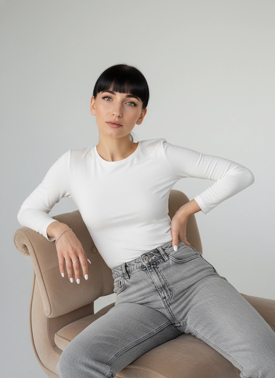 Роскошная уверенная женщина в минималистичной студии, смотрит прямо в камеру.
Одежда: белая блузка, плотно облегающая; светло-серые джинсы с высокой посадкой, слегка расстёгнуты.
Поза: женщина сидит верхом на дизайнерском бежевом кресле из вельвета, корпус развёрнут, одна рука лежит на спинке кресла, вторая — на талии, диагональ к лицу.
Волосы как на фото -убраны назад, чёлка как на фото.
Детали: белый маникюр, серебряное кольцо на безымянном, тонкий браслет.
Фон светлый, почти белый. Мягкий рассеянный студийный свет.
Фотореализм, высокая детализация фактур, плёночное зерно, 50mm, f/2.0–2.8.