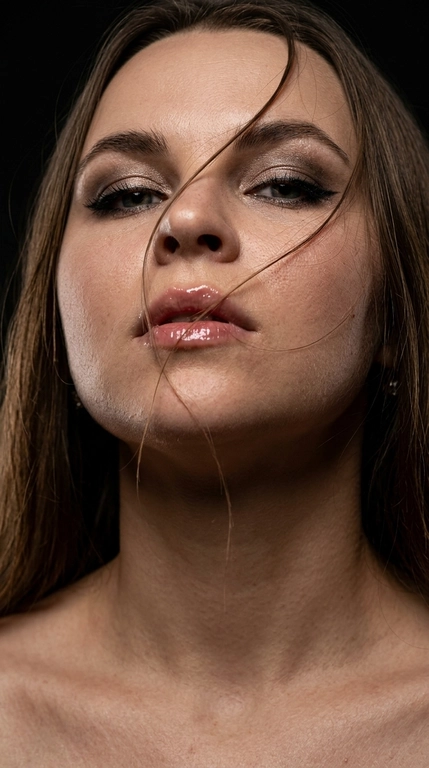 Фотореалистичный цветной beauty-портрет, экстремально крупный план (лицо + шея + ключицы), низкий ракурс снизу вверх, голова слегка запрокинута. Волосы хаотично пересекают лицо, несколько прядей на губах и щеке, ощущение ветра и движения. Взгляд открытый, дерзкий.
Макияж: дымчатые тени с мягким светлым шиммером, чёткая чёрная стрелка, густые ресницы, аккуратно оформленные брови. Губы, с влажным глянцевым блеском, чёткий контур. Кожа без ретуши — естественный чистый  тон, живая текстура, видимые поры, мягкие цветовые переходы света и тени.Модадвя девушка не меняй лицо 
Фон полностью чёрный, минимализм.
Свет: один боковой soft key light + лёгкий fill, высокий контраст, глубокие тени, выразительный рельеф скул и губ. Кинематографичный цветовой свет с естественными оттенками кожи и лёгкими тёпло-холодными нюансами. Лёгкий rim/back light для отделения от фона.
Съёмка в стиле fashion editorial. Сверхрезкий фокус на глазах и губах, малая глубина резкости. 
Ideal quality settings: ultra-realistic 8K, hyper-detailed, sharp focus; shallow depth of field, natural bokeh; HDR ,; cinematic color grading; lens 85mm / 50mm / 35mm; aperture f/1.4–f/2.8; key light at 45°, soft shadows; clean white balance; natural skin tones, glossy highlights.