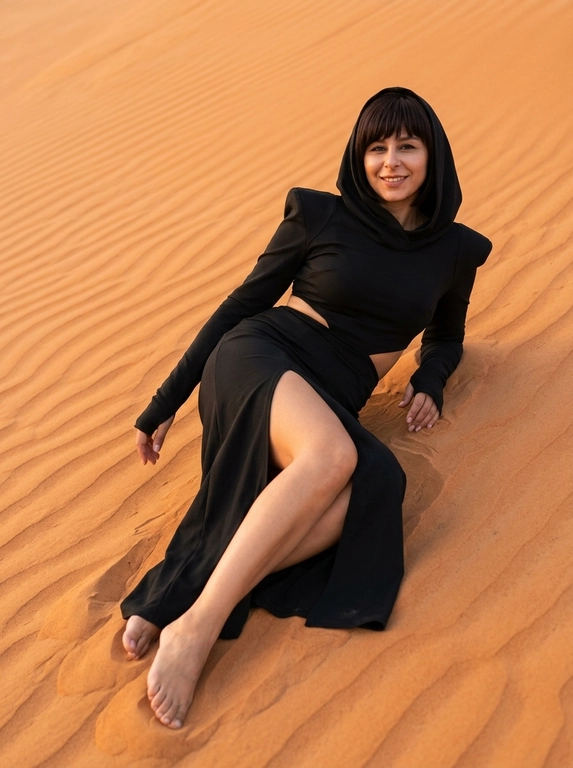 A hyper-realistic fashion photo of a woman reclining on the slope of a rippled orange sand dune during golden hour. Full-body shot, the entire body visible from head to toes, both feet fully visible, no cropping, low angle, her body placed diagonally across the dune, leaning back on one arm while the other hand rests naturally near her thigh. One leg is extended, the other slightly bent through the high slit of the skirt. She looks directly into the camera with a calm, confident expression. She wears a dramatic black hooded outfit with structured shoulders, fitted long sleeves with thumbholes, a waist cutout, and a long skirt with a high slit. Minimal dewy makeup, clean white manicure, natural skin texture, realistic pores, no plastic skin, no CGI. Warm golden-hour light, soft sun glow, gentle shadows, detailed sand texture, realistic fabric folds, cinematic but natural atmosphere. Shot on Canon EOS R, 24-70mm lens, sharp focus, ultra-realistic editorial photography.

Доп. характеристики: цвет волос и прическу не менять