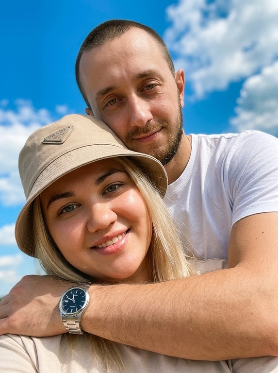 Кинематографичный кадр крупным планом (selfie style), пара в объятиях (классическое "обнимашки сзади") на фоне ярко-голубого неба с облаками.  Женщина на ней бежевая панама с брендовой надписью. Её кожа выглядит сияющей, подсвеченной мягким естественным солнечным светом. Выражение лица спокойное, глубокий взгляд, направленный прямо в камеру. Мужчина стоит строго позади девушки и обнимает её сзади, одна его рука обнимает её за плечи/шею спереди — предплечье лежит поперёк её ключиц, кисть свободно свисает с её плеча, на запястье классические серебряные часы с черным циферблатом. Мужчина в белой футболке, выражение лица доброжелательное, его взгляд прямо в объектив. Ракурс сверху, камера сфокусирована на лице девушки. Высокий контраст, насыщенные цвета, мягкая прорисовка деталей кожи. Эффект пленочной фотографии, легкое зерно, атмосфера летнего дня, эстетика социальных сетей. Телефона не видно в кадре.