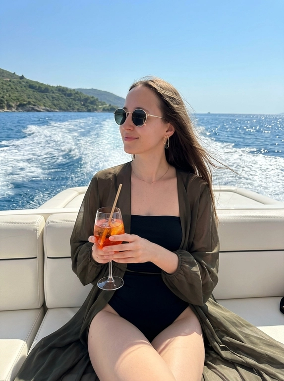 Фотореалистичное luxury lifestyle фото на борту скоростного катера. Кадр крупным планом, по пояс. Ракурс сверху, выше уровня глаз. Женщина сидит на белом кожаном сиденье катера, корпус слегка развернут в три четверти, голова повернута в сторону, взгляд направлен в сторону, спокойное расслабленное выражение лица, легкая полуулыбка. В одной руке бокал с ярко-оранжевым коктейлем со льдом и трубочкой, Минималистичные золотые серьги необычной формы, тонкая цепочка на шее. Волосы естественно развеваются ветром. Позади катера пенится кильватерный след, лазурная вода с солнечными бликами, зеленое побережье и невысокие холмы на горизонте, атмосфера дорогого летнего отдыха. Яркое полуденное солнце, чистое голубое небо, насыщенные естественные цвета, выразительные блики на воде и украшениях, дорогая цветокоррекция с пленочной эстетикой Kodak Gold 200, теплые оттенки кожи, глубокие синие и бирюзовые тона воды, естественный контраст без пересветов. Съемка на iPhone 16 Pro, 24 mm, photorealistic skin texture, luxury lifestyle photography, natural depth of field, ultra realistic, high detail, 8K.

Какая будет одежда?:  На ней черный купальный костюм без бретелей и легкая короткая полупрозрачная темно-оливковая шифоновая накидка, свободно накинутая на плечи. Тонкая ткань эффектно развивается сильным морским ветром, создавая красивые воздушные складки и динамику кадра. 

Очки:  круглые солнцезащитные очки в золотой оправе с темными линзами