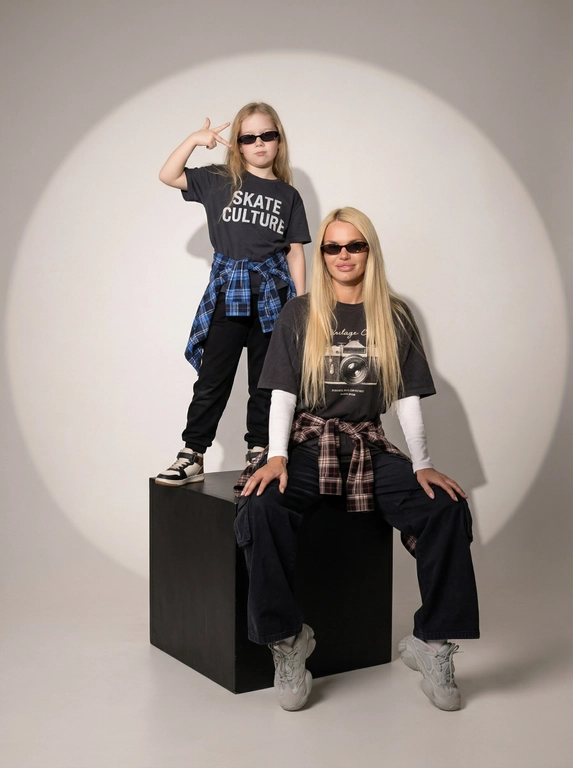 Photorealistic studio portrait photography in minimalistic fashion/editorial aesthetics, static camera at chest level of a seated person, light frontal angle without noticeable perspective deformation; composition with two characters: an model sits on a large matte black cube, body slightly turned towards the camera, knees bent, feet on the floor, the arms are lowered and relaxed in front of the knees, the fingers are neatly extended; the child stands on the same cube behind and to the left, at full height, one hand raised with a "V" gesture, the other lowered, pose confident and playful; both wearing narrow dark sunglasses, gaze directed at the camera; shooting plan — full height for both with a small supply of air above and below, center the center of the frame is shifted to the right to a sitting figure, a standing child balances on the left; the lens is normal/short portrait 50-70 mm, without width; aperture is approximately f/5.6–f/8, moderate depth of field, both characters are sharp, the background is slightly soft; the studio lighting is soft, with one key source on the front left at a level just above the head (softbox), giving smooth transitions and a pronounced but soft shadow against the background on the right; light filling light from the front reduces contrast, shadows are readable; the background is a smooth, light, seamless cyclorama/wall with a noticeable large oval light spot (gradient) in the center, there is a darker vignetting around, a clean minimalistic studio without props; the mood is calm, stylish, slightly retro, with an emphasis on street fashion and family vibe; the expression on the face of the seated model is a soft smile, relaxation; the person standing has a confident neutral facial expression; clothes: the seated model has a dark oversized T-shirt with a large rectangular print on the chest, under it a light longsleeve with long sleeves, wide dark cargo trousers/joggers with pleats at the knees and bottom, a plaid shirt/flannel tied at the waist with a knot, massive light sneakers with a pronounced sole and mesh texture; the child has a dark T-shirt with a large inscription on the chest, dark trousers, a plaid shirt tied at the waist, sneakers in two colors; fabrics with a realistic cotton and flannel texture, noticeable fine folds, natural creases on the knees and in the waist area, matte surfaces without gloss; micro-details: natural skin texture without plastic, neat glare on the lenses of glasses, a clear contour of the shadow from the raised hand on the background, clean floor and soft drop shadow from the cube; high detail, natural sharpness, light grain like a film shot, moderate contrast, soft matte/film look, no HDR, no overexposure and no unnecessary objects.

Какого цвета будут рубашки на поясе?: Красный и синий.