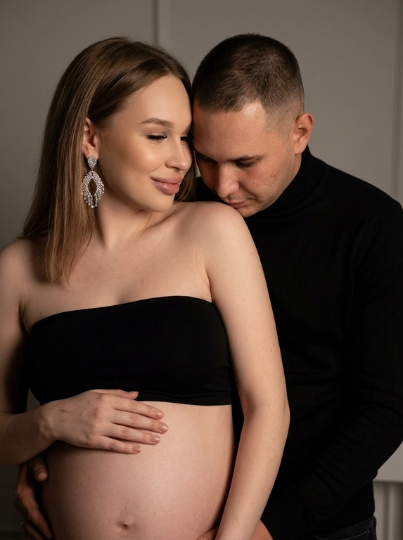 Интимная стильная сцена с трогательной эмоциональной связью. Женщина в полупрофиле к камере с легкой улыбкой, в минималистичном черном топе без бретелек, показывающий открытый беременный живот с мягкой матовой кожей под равномерным светом, руки на животе, создавая заботу. Крупные висячие серьги с видимой фактурой и блеском. Мужчина позади, наклонив голову вниз к ней, в черном тонком свитере с высоким воротом, подчеркивающем стройность. Мягкое освещение, серая фональная поверхность, тени подчеркивают рельефы и создают игру света и тени, как тихий вечер в закрытом пространстве. Композиция: мужчина словно обнимает женщину, руки частично обрезаны. Лицевые черты сохранены, передавая индивидуальность и естественную красоту, атмосфера любви, ожидания и легкой загадочности. Фотореалистично, высокая текстурная детализация, 8K.