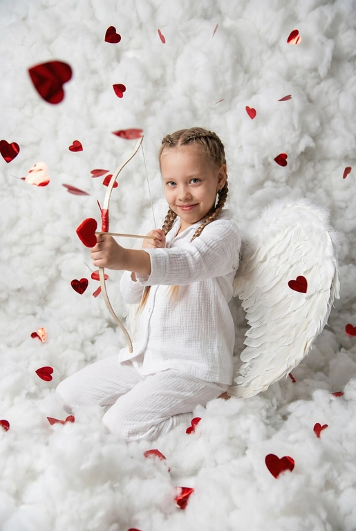 Использовать реальные черты лица человека с референса, естественную текстуру кожи и настоящий цвет и длину волос. Личность должна оставаться неизменной.

Y2K. Сказочная сцена с купидоном, сфотографированная как реальный момент.
Человек сидит среди плотных, пушистых белых облаков, которые полностью заполняют передний, средний и задний план, образуя бесшовную белую среду с многослойной глубиной.

Поза вполоборота: корпус слегка повернут вбок, одно плечо выдвинуто вперед, как будто человек натягивает и отпускает небольшой лук с красной стрелой в форме сердца; руки подняты с легким напряжением, указывающим на движение при выстреле.

Несмотря на поворот тела, голова повернута к объективу, взгляд направлен прямо в камеру. Выражение лица радостное и игривое, с мягкой улыбкой.

Наряд — простая белая пижама: свободная рубашка и брюки из муслина.
На спине — крылья из белых перьев, настоящие и фактурные, с четкими бликами по краям каждого пера.

Вокруг объекта в воздухе хаотично летают только конфетти в форме сердечек — маленькие красные сердечки из глянцевой фольги, расположенные на разном расстоянии: часть в переднем плане, часть растворяется среди облаков.

Без рассеянного света, без мягкого свечения, без студийного глоу.
Текстура кожи реалистичная и детализированная, без сглаживания и без фильтров.
Тонкая зернистость, волшебное, но искреннее ощущение Y2K-редакции — выглядит как фотография, а не визуализация.