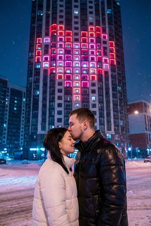  Создай не меняя черты лица. Романтический снимок пары, стоящей на заснеженной городской улице поздним вечером. Главный объект – крупный план пары: мужчина в черной кожаной дутой зимней куртке нежно целует женщину в белой дутой куртке в лоб, она стоит с прикрытыми глазами, улыбаясь. За ними возвышается массивный современный жилой небоскреб, на фасаде которого окна на нескольких этажах подсвечены ярким  светом, формируя огромное пиксельное сердце. Освещение драматичное, контрастное, с преобладанием холодных тонов в небе и тёплого, неонового розового свечения от здания. Снег на земле отражает свет. Урбанистическая, зимняя, кинематографическая фотография, атмосферный кадр.