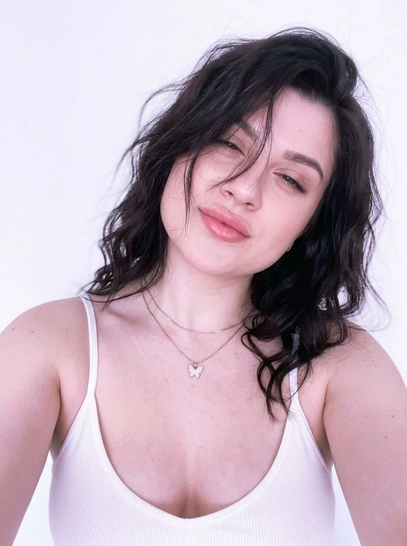 Vertical close-up portrait in the style of a natural selfie: a woman with her head tilted and a soft playful expression, one eye squinted or closed, gaze directed at the camera.
Expressive full lips, neat eyebrows, and soft makeup with an emphasis on the lips and eyes are noticeable on the face. There are thin chains around her neck, one with a small butterfly pendant.
The clothes are a white ribbed tank top with thin straps, revealing the shoulders and upper chest. The background is almost completely white, empty and minimalistic, like a light wall, without unnecessary objects.
The light is very soft, diffused, slightly overexposed, with a cool pink and purple hue, creating a gentle, slightly dreamy atmosphere.
The composition is close and intimate, the frame is cropped around the shoulders and chest, with emphasis on the face. The camera is at face level, creating the feeling of taking a picture from a phone. Light graininess, a gentle loss of detail in the highlights, natural, fashionable, slightly vintage aesthetic.



Причёска: Wavy hair, slightly tousled, with loose strands naturally falling across parts of the face without covering the eyes.