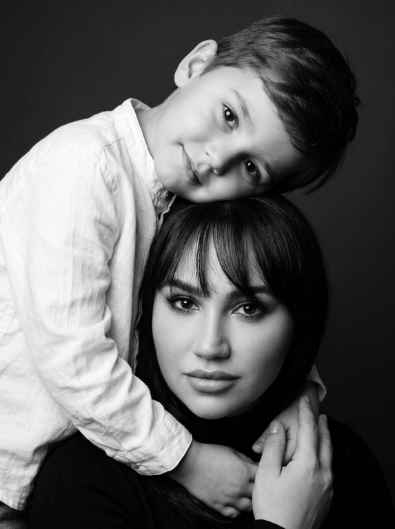 Родитель и ребёнок с внешностью как на приложенных фото, художественная семейная студийная фотосессия. Композиция — вертикальный портрет, крупный план лиц. Родитель внизу смотрит прямо в камеру, лицо фронтально. Ребёнок сверху, голова повернута набок, щека лежит на голове родителя, смотрит в камеру с нежной улыбкой. Рука ребёнка обнимает родителя за шею, видна в кадре, родитель держит руку ребёнка своей рукой.
Одежда: ребёнок в белой льняной рубашке, родитель в чёрной водолазке. 
Фон — тёмно-серый почти чёрный однотонный студийный, создаёт контраст. Освещение — мягкое боковое студийное, создаёт объём и глубину.
Чёрно-белая художественная фотография с выразительными контрастами. Атмосфера нежная, интимная, спокойная.