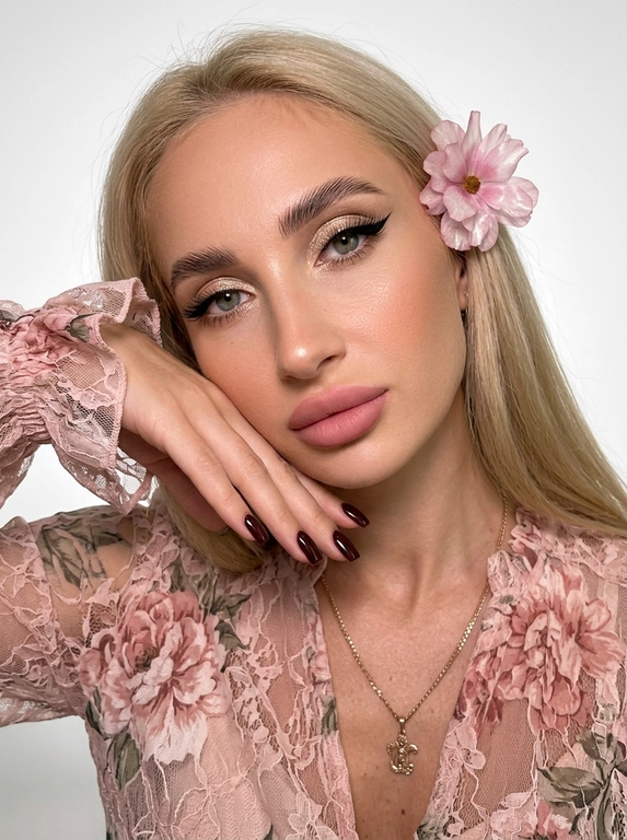 На фотографии запечатлена женщина крупным планом, позирующая для портрета. Снимок подчеркивает её ухоженную внешность и стильный образ.
Лицо и выражение:
•  Глаза:  ярко выделены макияжем. На верхних веках нанесены мерцающие золотисто-бежевые тени, дополненные четкими, длинными черными стрелками («кошачий глаз»). Ресницы густые и длинные, подчеркивающие глубину взгляда.
•  Брови: Густые, уложенные вверх (эффект ламинирования), что придает лицу современный и открытый вид.
•  Кожа: Тон кожи ровный, с нежным персиковым румянцем на скулах. Кончик носа и скулы слегка подсвечены хайлайтером для придания лицу сияния.
•  Губы: Выразительные, накрашены матовой помадой нежно-розового оттенка. Верхняя губа имеет четкий контур.
•  Поза: Женщина слегка наклонила голову вправо и подперла щеку правой рукой. Этот жест придает снимку нотку нежности и кокетства.Маникюр темно-бордовый,квадратный.
Одежда:
•  Ткань украшена принтом в крупный кружевной цветок .
•  Рукава имеют пышные воланы или оборки у запястий, что добавляет образу романтичности и женственности.
Фон и освещение:
•  Фон белый студийный
•  Освещение мягкое, направленное на лицо, что позволяет рассмотреть все детали макияжа и создает приятную, эстетичную картинку.
Общее впечатление:
Фотография представляет собой классический бьюти-портрет. Образ женщины сочетает в себе элегантность, современность и романтичность. Тщательно проработанный макияж и стильная одежда создают гармоничный и привлекательный облик.

Какого цвета будет платье?: На женщине надета блузка или платье из легкой, полупрозрачной ткани нежно розового цвета.

Какого цвета будет цветок?: В волосах один нежный ,воздушный розовый цветок.