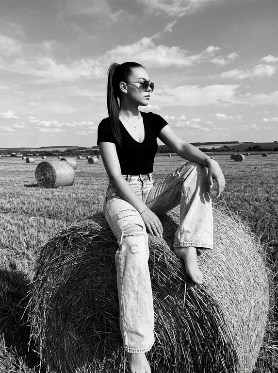 Чёрно-белая high-fashion editorial фотография в сельской местности, женщина с внешностью как на референсе, черты лица не изменять, композиция — более общий план (wide shot), камера на среднем расстоянии, фигура полностью в кадре с окружающим пространством, она сидит на большом тюке сена в поле, корпус слегка развернут, одна нога свободно опущена вниз, вторая согнута, создавая расслабленную, но выразительную позу, руки естественно лежат на колене и на поверхности тюка, взгляд направлен в сторону, профиль, выражение спокойное и уверенное, одежда: чёрный топ с V-образным вырезом и объёмные светлые широкие джинсы с потёртостями и рваными элементами, аксессуары: крупные солнцезащитные очки, минималистичные украшения, фон: открытое поле с несколькими тюками сена на разной глубине, создающими перспективу, линия горизонта ниже центра кадра, чтобы подчеркнуть небо, освещение: жёсткий естественный дневной свет с мягкими облаками, формирует выразительные тени и контраст, стиль: кинематографический, плёночное зерно, высокая детализация, реалистичная текстура кожи и ткани, редакционный fashion стиль, акцент на атмосфере и пространстве кадра


Какая будет прическа?: Гладкие волосы собраны в высокий хвост