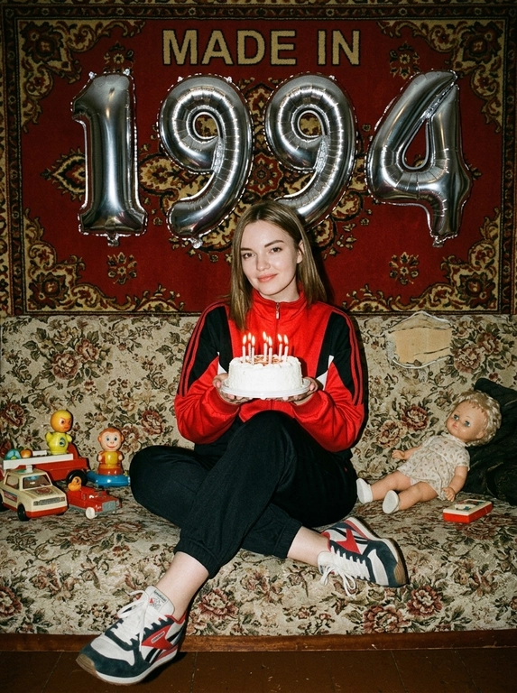 Создай крупный портрет, не меняя черты лица. На фотографии модель в стиле «лихие 90-е» в России. Модель сидит на старом диване с цветочной обивкой на фоне красного советского ковра на стене и смотрит в камеру с лёгкой улыбкой. На ковре висят большие надувные серебряные фольгированные шары в форме цифр и надпись «MADE IN». На модели классический красный спортивный костюм того времени и кроссовки. В руках маленький праздничный белый торт со свечами. Рядом на диване лежат игрушки: советские игрушки и старая советская кукла. Фото со вспышкой. Освещение мягкое, создающее атмосферу ностальгии. Высокое разрешение, реализм. Модель в олдскульном спортивном костюме сидит расслабленно, с лёгкой улыбкой. Ретро-вайб, приглушённые цвета, зернистость плёнки, детализированные текстуры ткани и шерстяного ковра.

Какая будет цифра?: Цифра 1994.