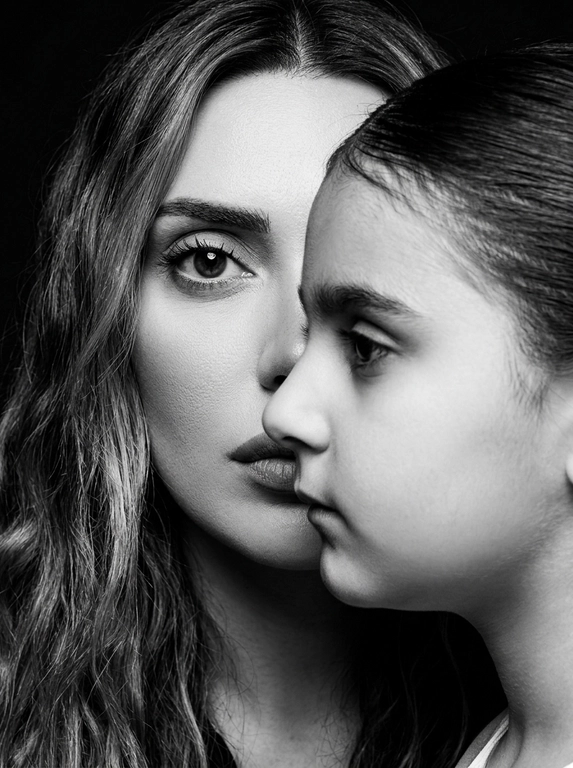 Чёрно-белый фотореалистичный студийный beauty-fashion портрет родителя и ребёнка с представленных фото. Крупный плотный кадр по плечи, без свободного пространства. Ребёнок находится на переднем плане, ближе к камере, в профиль, расположен сбоку и частично перекрывает лицо родителя. Лицо ребёнка закрывает примерно половину лица родителя, создавая сильный эффект визуального перекрытия. Родитель находится позади ребёнка, строго по центру кадра; у родителя виден только один глаз, направленный прямо в камеру, вторая часть лица скрыта лицом ребёнка. Лица расположены максимально близко, почти соприкасаются, но без явного физического действия. Ребёнок смотрит в сторону, выражение спокойное, сдержанное и закрытое. Родитель смотрит прямо в камеру одним открытым глазом, взгляд холодный, спокойный и пронизывающий. Атмосфера high-fashion editorial: минимальная эмоциональность, люксовая отстранённость, скрытое напряжение и холодная близость. Фон тёмный, чистый, студийный, без деталей. Жёсткое студийное освещение, высокий контраст, глубокие тени, выразительная светотень на лицах, натуральная текстура кожи, sharp focus, dramatic black and white photography, editorial beauty portrait, без улыбок, без лишних предметов, без текста и логотипов.