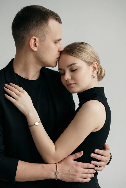   Мужчина и женщина позируют в студии в объятиях. Мужчина стоит позади женщины, обнимая её обеими руками; его тело повернуто к ней, голова слегка наклонена вниз, губы касаются её лба. Женщина немного отклоняет голову назад к его плечу, её глаза закрыты. Кадр — средний, от талии до головы, снят на уровне глаз. Композиция плотная, акцентирует линии рук и соприкосновение лиц. Женщина стоит, прямо обращённая к камере.
На ней облегающий чёрный топ без рукавов с высоким воротом, из плотной матовой ткани.
Мужчина одет в чёрную футболку и чёрную неброскую куртку. На обоих запястьях — тонкие металлические браслеты с минималистичными круглыми подвесками, а женщина носит небольшие светлые металлические серьги с гладкой поверхностью.
Обстановка минималистичная: однотонный светло-серый фон без текстуры.
Освещение — мягкий студийный свет: рассеянный фронтальный и боковой свет создаёт плавные переходы света и тени на лицах и руках. Тени мягкие, средней глубины. Контраст умеренный, без жёстких бликов. Свет равномерно распределён по фигурам, фон подсвечен мягко, без выраженных градиентов.
Съёмка на портретный объектив 85 мм, малая глубина резкости с чётким фокусом на лицах и руках; фон слегка размыт, текстуры кожи и ткани высоко детализированы. Это минималистичный студийный портрет, высокодетализированный и фотореалистичный.
Минималистичное романтическое студийное настроение, нейтральные тёплые тона кожи, низкая–средняя насыщенность, мягкое рассеянное освещение, средний контраст с нежными тенями, плавные переходы бликов, естественная передача кожи, чистый светлый фон, тонкий тональный баланс, современная мягкая редакционная цветокоррекция. Волосы женщины собраны в низкий, гладкий пучок с аккуратно уложенными прядями.