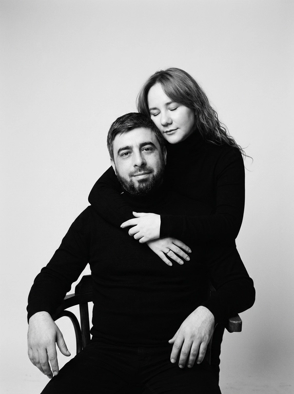 Черно-белая студийная портретная фотография в стиле минимализма. На снимке изображены мужчина и женщина, позирующие на нейтральном светлом фоне. Мужчина сидит на стуле лицом к камере, его руки расслабленно лежат на подлокотниках, взгляд направлен прямо перед собой с мягкой полуулыбкой. Женщина стоит сзади, нежно обнимая мужчину за грудь руками, а ее голова аккуратно прислонена щекой к макушке мужчины с закрытыми глазами. Оба одеты в одинаковые черные водолазки с длинными рукавами. Атмосфера кадра интимная, теплая, спокойная и эмоционально близкая. Цветовая гамма монохромная, выдержанная в контрастных черно-серых тонах с мягким студийным освещением, подчеркивающим текстуру одежды и контуры лиц.