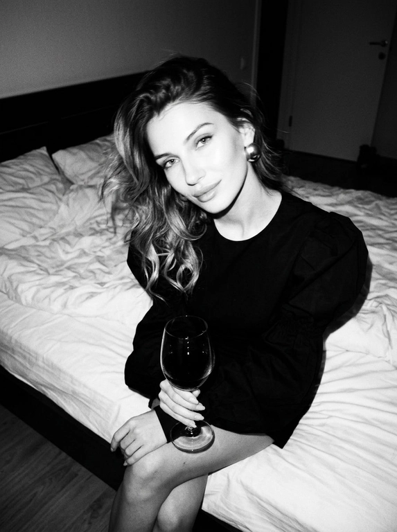 Create a portrait, transfer the face from the reference photo without changing facial features, proportions, or expression, preserve full identity. Aesthetic black-and-white nighttime Y2K photograph captured with direct on-camera flash, shot in motion.
A woman is sitting on the edge of a white, slightly rumpled bed, the frame taken from a slightly elevated angle. She wears a stylish black top with voluminous sleeves and large round statement earrings. In one hand she holds an elegant glass of dark wine. Her body is subtly moving, creating natural motion blur across the face and shoulders, as if the moment was caught mid-gesture. The gaze is languid and directed toward the camera, with a soft, intimate smile. Hair falls in large waves with loose strands drifting across the face, slightly displaced by movement. Lighting is harsh flash against deep night shadows, high contrast with blown highlights and strong falloff, visible film grain. Skin texture remains real and alive, no smoothing, enhanced by flash glare. The mood is high-fashion editorial, Parisian chic, old-money sensuality, nighttime melancholy, spontaneous and cinematic, shot in a raw 35mm film aesthetic with pronounced motion blur and lived-in realism.