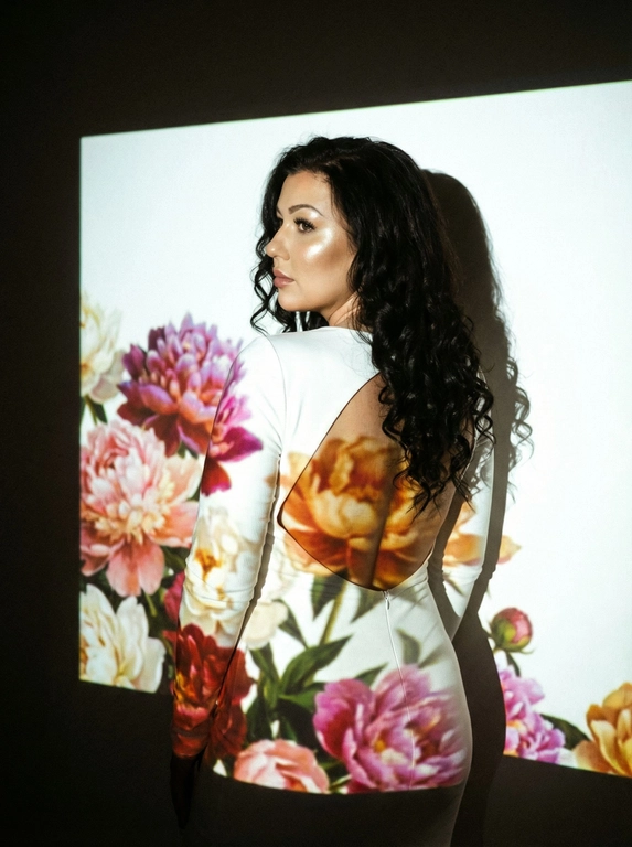 Кинематографический эффект с добавлением зерна. Ультрареалистичный портрет женщины, стоящей на фоне белой стены: её тело немного повёрнуто в сторону.  Фото с нижнего ракурса, фото со спины девушки. Макияж лёгкий: хайлайтер, длинные ресницы. На ней белое однотонное платье по фигуре, с открытой спиной и длинными рукавами. Она не смотрит в камеру. Освещение мягкое, с лёгким акцентом на её лицо, создающее нежные тени и подчёркивающее черты. Вокруг темно — светит только проектор с изображением ярких пионов прямо на девушку и на стену за ней. Только один источник света — проектор.


Какая будет прическа?: Длинные волосы, собранные в мягкие локоны