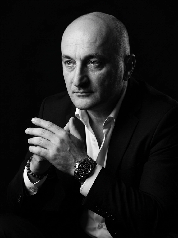 A hyper-realistic 8K cinematic portrait of a uploaded person,
captured in a dramatic Chiaroscuro style. The subject is seated at a
three-quarter angle, leaning slightly forward with a relaxed yet
commanding posture. His face is turned away from the lens, with
one side of his face clearly illuminated while the other is
completely lost in deep, velvety black shadows. His expression is
contemplative, not looking at the camera. The pose is precise: his
hands are clasped together near his chest; one hand is held
straight with fingers slightly tilted, while the other hand is
positioned so only the fingers are visible, resting over the first. He
wears a luxury black chronograph watch with a detailed metal link
bracelet, and a beaded bracelet. His attire is a sharp
black suit jacket over a white dress shirt with the top buttons
open, showing refined fabric textures. The background is a solid,
seamless black. The lighting is sharp and directional, highlighting
the intricate details of his hair,
the moisture in his eyes, and the glint off the watch face.
Cinematic black-and-white color grade with rich contrast and
smooth, natural skin transitions. Shot on 85mm lens, f/1.8, high-
resolution commercial photography