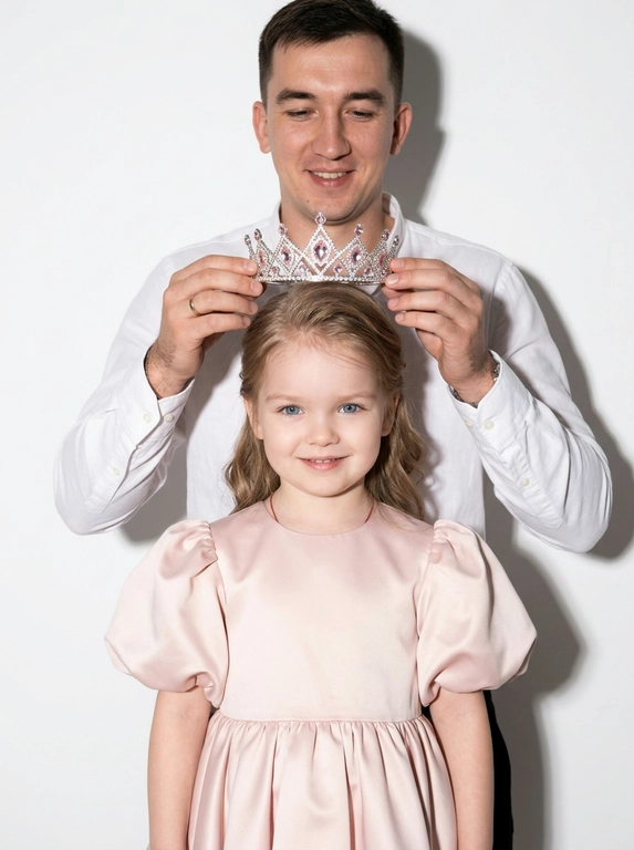 Use the uploaded reference photo and preserve the facial features, bone structure, skin tone, and identity exactly 100% identical to the original. Do not change the face shape, individuality, or expression. Maintain realistic skin texture with visible pores and natural proportions.Create a color studio portrait. The photo is shot in darkness with a strong direct on-camera flash against a clean white background, creating crisp highlights and defined shadows while the surrounding space remains dark. Close portrait composition focused on a man and the child from the photo.The child from the photo stands in the foreground looking toward the camera with a soft gentle smile. The man stands behind slightly higher in the frame, smiling softly while holding an elegant silver crown decorated with delicate light-pink diamonds just above the child’s head, in the moment of placing it on — the crown is not yet on the head.The man wears a white shirt with the top buttons open at the neck. The child wears a luxurious soft light-pink satin dress, only the upper part visible in the frame, with voluminous puff sleeves and structured shoulders. Hair styled brushed back into soft waves.Ultra-realistic detail, natural skin texture, cinematic flash photography, clean white studio background, elegant intimate atmosphere.