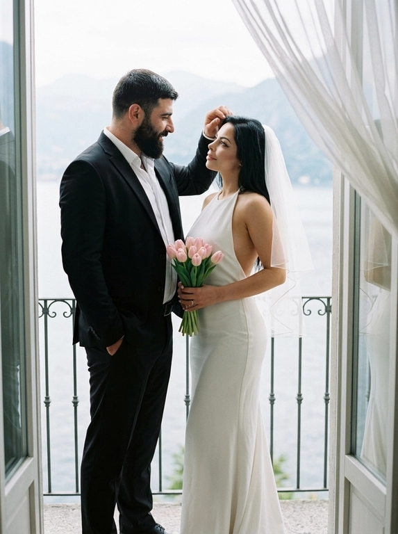 Фотореалистичный bridal editorial, полный рост. Пара стоит на узком балконе у открытой стеклянной двери. Мужчина слева, корпус в 3/4 вправо к женщине, одна рука в кармане брюк, другой рукой мягко касается её волос у лба, голова чуть наклонена к ней, взгляд на женщину, лёгкая нежная улыбка. Женщина справа, стоит очень близко, корпус в 3/4 влево, голова поднята к мужчине, взгляд снизу вверх на его лицо. В обеих руках прижат к животу маленький аккуратный букет из 11 этих цветов: бутоны цветов собраны плотно, стебли строго вниз, букет вертикальный, не у бедра, не у груди. Мужчина в чёрном костюме и белой рубашке без галстука. Женщина в длинном белом платье-халтер с открытой спиной. Фон: кованые перила, озеро, горы, светлые шторы. Мягкий естественный дневной свет, 50mm, лёгкое зерно.

Из каких цветов будет букет невесты?: Нежно-розовые тюльпаны

Доп. характеристики: На волосах фата у девушки