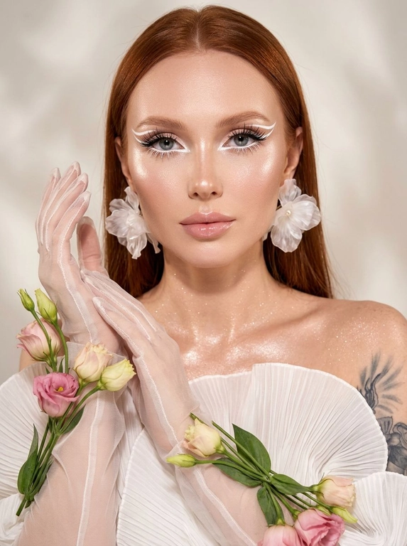 Создай крупный портрет в стиле высокой моды, не меняя черты лица. Элегантный макияж глаз с длинными острыми белыми двойными изогнутыми стрелками, длинные густые ресницы эффект лучики, нижние густые ресницы накрашены белым, белый каял карандаш на нижнем веке, белый хайлайтер во внутренних уголках и на скулах. Тело и декольте подсвечены хайлайтером и шиммером. На ушах объемные полупрозрачные белые серьги-цветы. Высокая детализация текстур кожи и одежды. Лицо освещено ярким белым светом, изящный кадр с нежным светом, мягко обволакивающим фигуру и подчеркивающим пышные складки белого платья из тонкой плиссированной ткани, создающей легкость. В длинных полупрозрачных перчатках, добавляющих изысканности и загадочности. По запястьям из-под перчаток и по предплечьям живые цветы — розовые и кремовые бутоны эустомы и листья, контрастирующие с кожей и блеском перчаток. Лицо обращено к камере, выражение спокойствия и мечтательности, с влажной сияющей кожей. Равномерно светлый нейтральный фон акцентирует фигуру и детали, тени размыты, создавая атмосферу тепла и уюта. Композиция сосредоточена на верхней части тела и руках, подчеркивая грациозность и изящество, фотореалистичное изображение, высокая детализация, 8K, высокая контрастность. Девушка смотрит прямо в камеру.