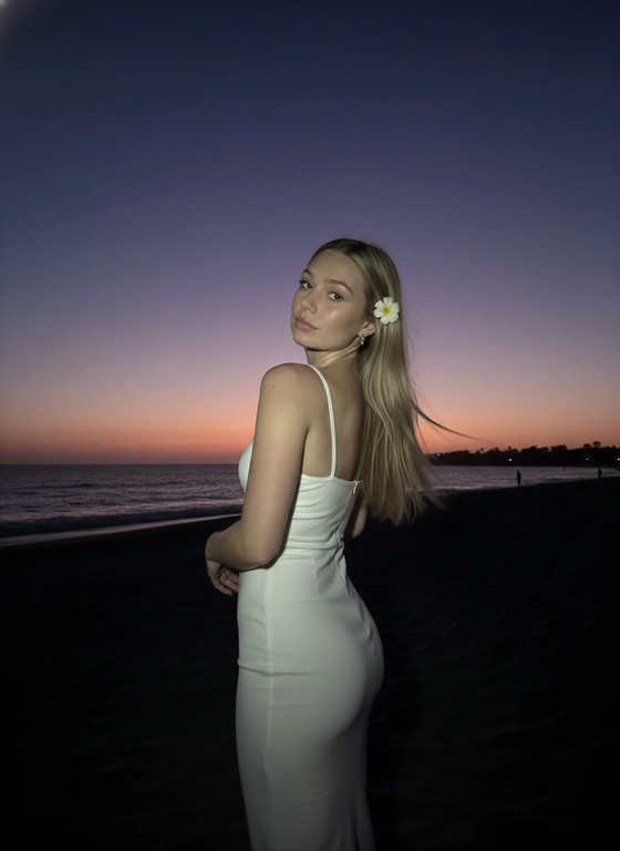 Сделай фото, будто я на пляже, где темно, но кто-то сфотографировал меня со вспышкой, и мое лицо немного блестит и светится, заднем плане закат, как будто солнце село на 98%, оставив фиолетовый оттенок, и само по себе почти темно. Фото сделано из-за моей спины, как будто  мне сказали, повернуться. Я в белом облегающем платье с цветком, заправленным в волосы. Волосы должны развеваться так, как будто дует слегка ветер.
