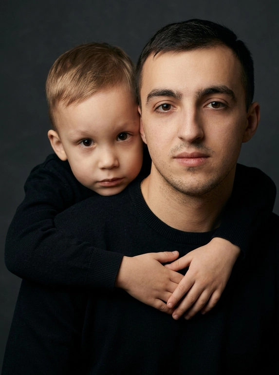 Создай портретный студийный кадр из фотосессии отца и сына. Сын обнимает отца сзади, лежа подбородком на его плече, руки мягко сомкнуты на груди мужчины. Оба смотрят прямо в камеру. Естественные спокойные эмоции, лёгкая серьёзность во взгляде.
Одежда — однотонные чёрные лонгсливы или свитера без принтов. Тёмный нейтральный фон (глубокий серо-оливковый или графитовый). Мягкий направленный студийный свет с лёгкой теневой глубиной, акцент на лицах, тёплая естественная цветокоррекция, высокая детализация кожи, натуральная текстура без излишней ретуши.
Кадр — крупный или средний план, плотная композиция, лица расположены близко друг к другу. Атмосфера — тёплая, камерная, эмоциональная, ощущение близости и доверия. Профессиональная съёмка, 85mm lens, shallow depth of field, cinematic portrait, ultra realistic.
