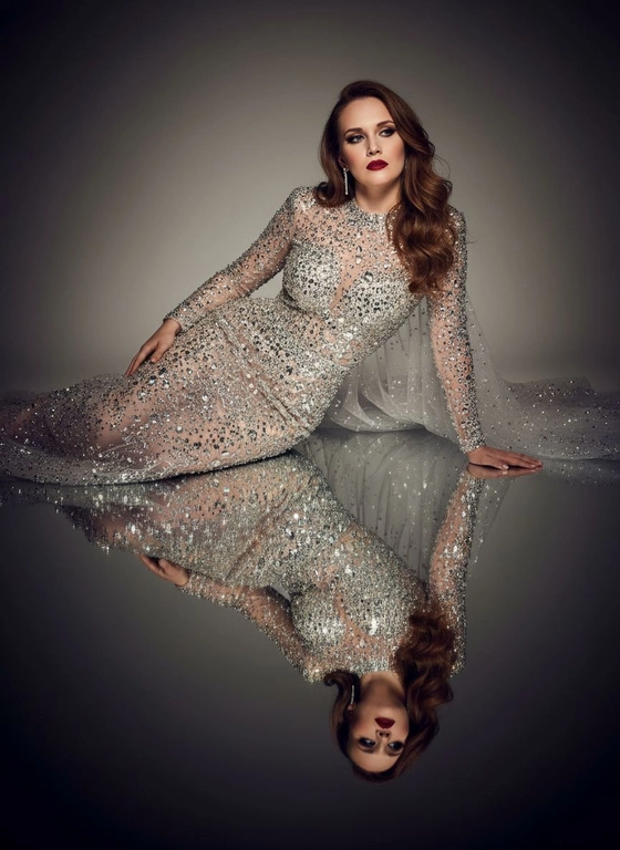 Это изысканный портрет женщины, лежащей боком на зеркальной поверхности, создавая идеальное отражение всего ее тела. На ней элегантное, облегающее платье от кутюр, полностью расшитое серебряными стразами, кристаллами и сверкающими деталями, которые роскошно ловят свет. Платье имеет длинные рукава, высокий ворот и струящийся шлейф из прозрачного тюля, усыпанного стразами на юбке, источающий гламур и изысканность. У нее длинные, волнистые волосы, которые нежно падают на ее плечи. Ее макияж безупречен с яркими темными smoky eyes и черной подводкой для глаз, длинными, объемными ресницами, безупречной кожей и пухлыми губами с изысканной, яркой красной помадой. На ней сдержанные, но элегантные серьги, дополняющие гламурный образ. Сцена драматически освещена темной виньеткой, подчеркивающей блеск платья и контраст между ее телом и отражением в зеркале. Минималистичный фон в нейтральных тонах ещё больше подчёркивает харизму модели и богатство деталей наряда. Общая атмосфера создаёт ощущение роскоши, элегантности и изысканности, отражая моду и высокую моду. Сохранены те же черты лица без каких-либо изменений, тот же тон кожи и цвет волос. Качественный маникюр, профессиональное студийное качество изображения.