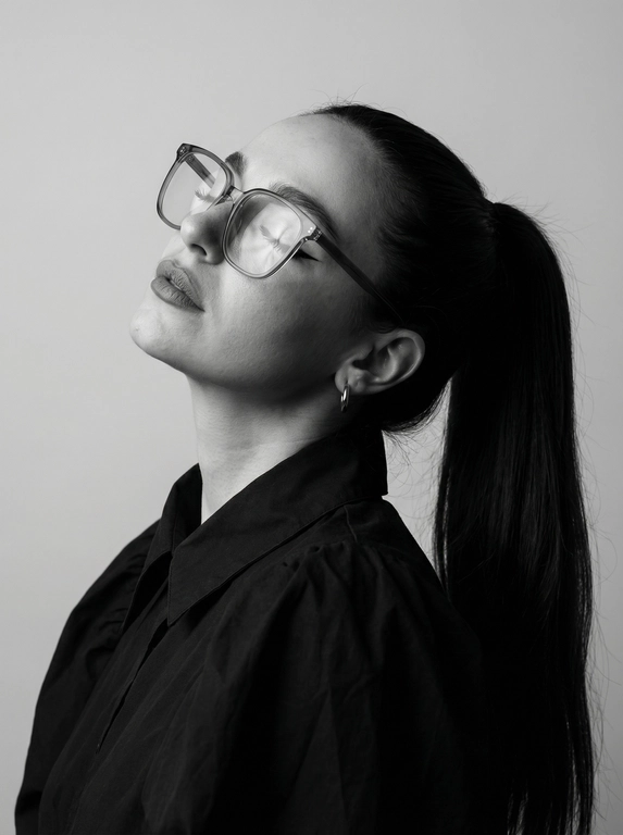 Чёрно-белый минималистичный фотореалистичный портрет, студийный мягкий свет спереди‑сверху, нейтральная светлая однотонная стена как фон. Девушка в профиль: голова запрокинута назад, подбородок поднят, глаза закрыты — расслабленная fashion-поза с акцентом на линии шеи и контуры лица. Обязательно точно сохранить черты лица с референс-фото (без искажений); крупные квадратные очки в массивной полу-прозрачной оправе с естественными бликами. Детали лица: длинные ресницы и чёткие стрелки, выразительные губы, естественная текстура кожи, аккуратный ретуш. Одежда: тёмный объёмный верх с выразительным воротником, маленькая серьга-кольцо. Стиль: высокая графичность и контраст, чистые линии, лёгкая драматичность, максимальная детализация и фотореализм, аккуратные светотеневые переходы, подчёркнутые контуры.

Какая будет прическа?: Гладкие волосы, высокий длинный густой хвост.